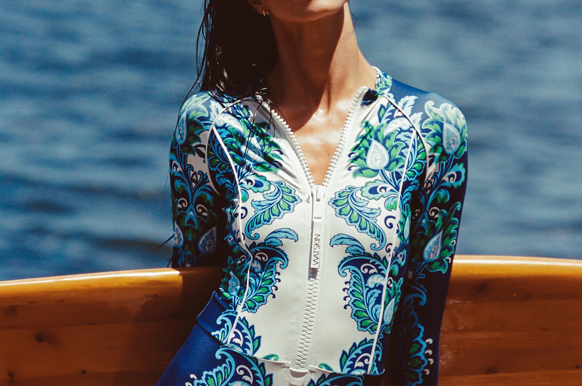 Woman in a blue and white patterned swimsuit standing by a wooden surfboard with ocean in the background