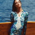Woman in a blue and white patterned swimsuit standing by a wooden surfboard with ocean in the background