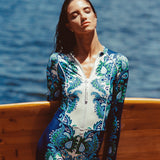 Woman in a blue and white patterned swimsuit standing by a wooden surfboard with ocean in the background