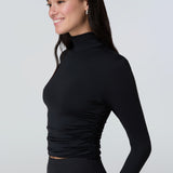 3/4 view of a woman wearing a black long-sleeve turtle neck rash-guard and bikini bottom.