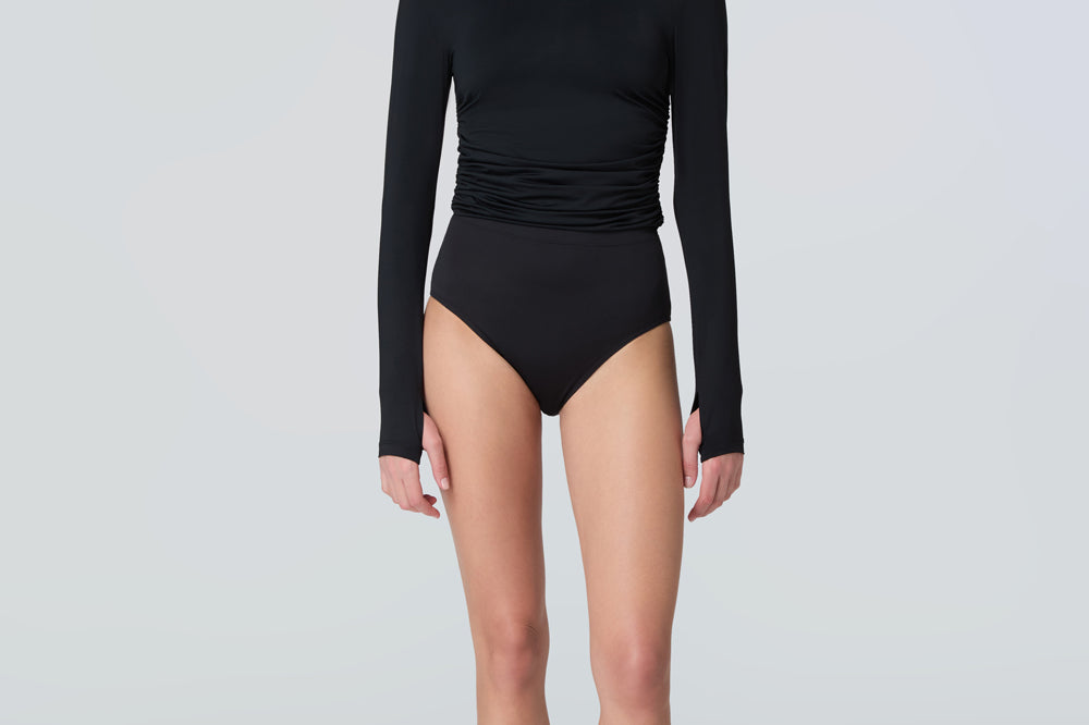 Full body view of a woman wearing a black long-sleeve turtle neck rash-guard and bikini bottom.