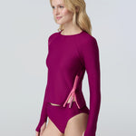 A woman wearing a long sleeve swim top in dark berry with contrasting pink zipper and dark berry swim bikini bottom.