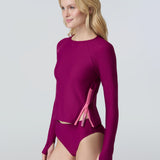 A woman wearing a long sleeve swim top in dark berry with contrasting pink zipper and dark berry swim bikini bottom.