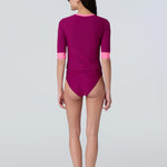 A woman wearing a short sleeve swim top in dark berry with contrasting pink trim and dark berry swim bikini bottom.