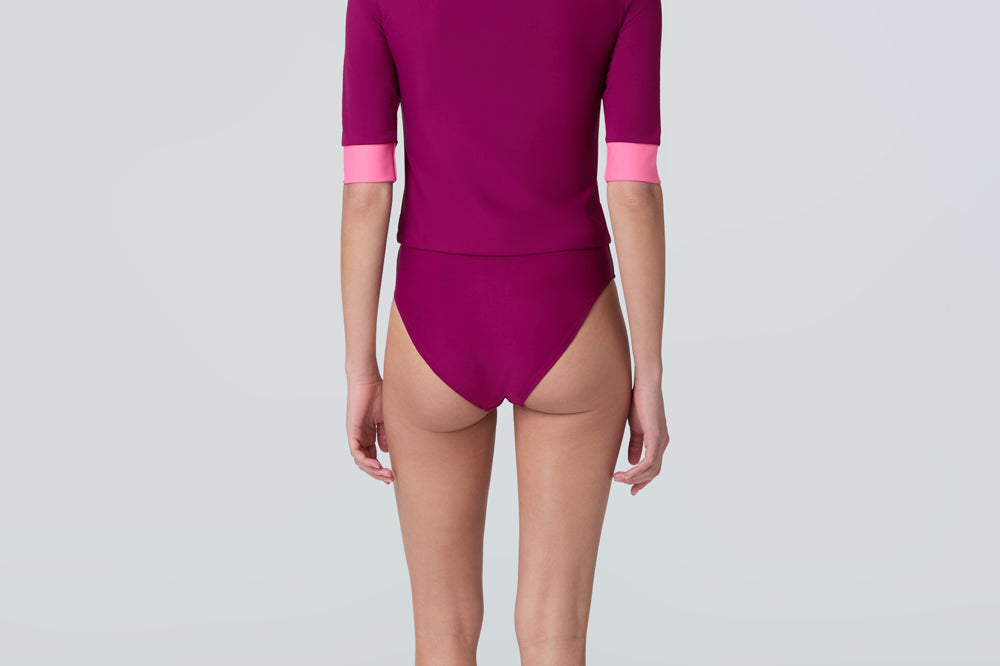 A woman wearing a short sleeve swim top in dark berry with contrasting pink trim and dark berry swim bikini bottom.