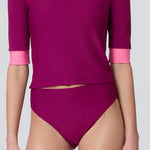 A woman wearing a short sleeve swim top in dark berry with contrasting pink trim and dark berry swim bikini bottom.