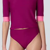 A woman wearing a short sleeve swim top in dark berry with contrasting pink trim and dark berry swim bikini bottom.