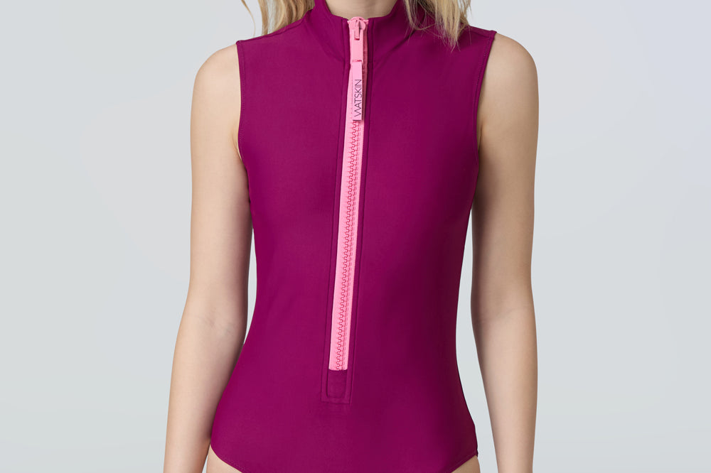 Front view of a woman wearing a sleeveless bodysuit in dark berry with pink zipper.