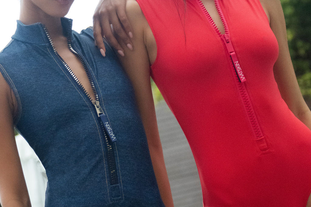 Two women modeling sleeveless one-piece swimsuits with front zippers in red and denim.