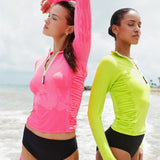 Two women at the beach wearing long-sleeve 1/2 zip rashguard with side ruching in lime green and pink with black bikini bottoms