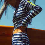 Woman wearing a blue & white striped long sleeve tee and boy short holding a surfboard in a tropical background.
