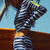 Woman wearing a blue & white striped long sleeve tee and boy short holding a surfboard in a tropical background.
