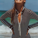 Woman in white swim goggles standing oceanfront modeling Watskin Lilia bodysuit in black/ivory houndstooth 