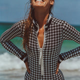 Woman in white swim goggles standing oceanfront modeling Watskin Lilia bodysuit in black/ivory houndstooth 