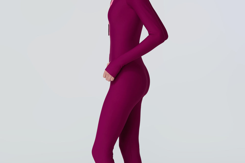 Side view of a woman wearing a long sleeve full length catsuit in dark berry with pink front zipper.