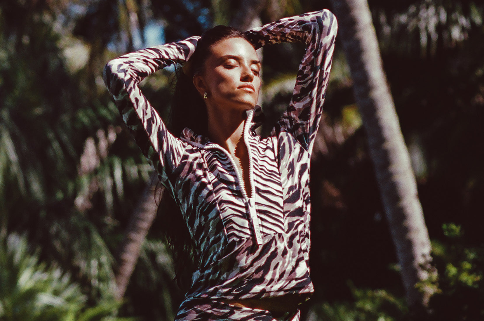 Woman modeling Watskin Mia half-zip rashguard and Aria high waist belted bikini bottoms in Animal Mix against a n outdoor tropical beach scene with palm trees