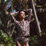 Woman modeling Watskin Mia half-zip rashguard and Aria high waist belted bikini bottoms in Animal Mix against a n outdoor tropical beach scene with palm trees