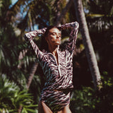 Woman modeling Watskin Mia half-zip rashguard and Aria high waist belted bikini bottoms in Animal Mix against a n outdoor tropical beach scene with palm trees