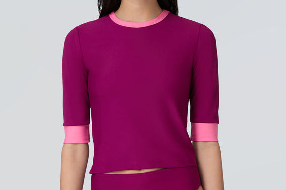 A woman wearing a short sleeve swim top in dark berry with contrasting pink trim and dark berry swim bikini bottom.