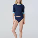 Front view of a woman wearing a navy blue short-sleeve top with light blue cuffs and neck trim and navy bikini bottom.