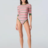 Full body front view of a woman wearing a pink and green wavy striped patterned short sleeve swim top with green sleeve cuff and bikini bottom with green trim on a white background