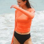 A woman wearing an orange long-sleeve rashguard with a zip sleeve pocket and side ruching and a black bikini bottom