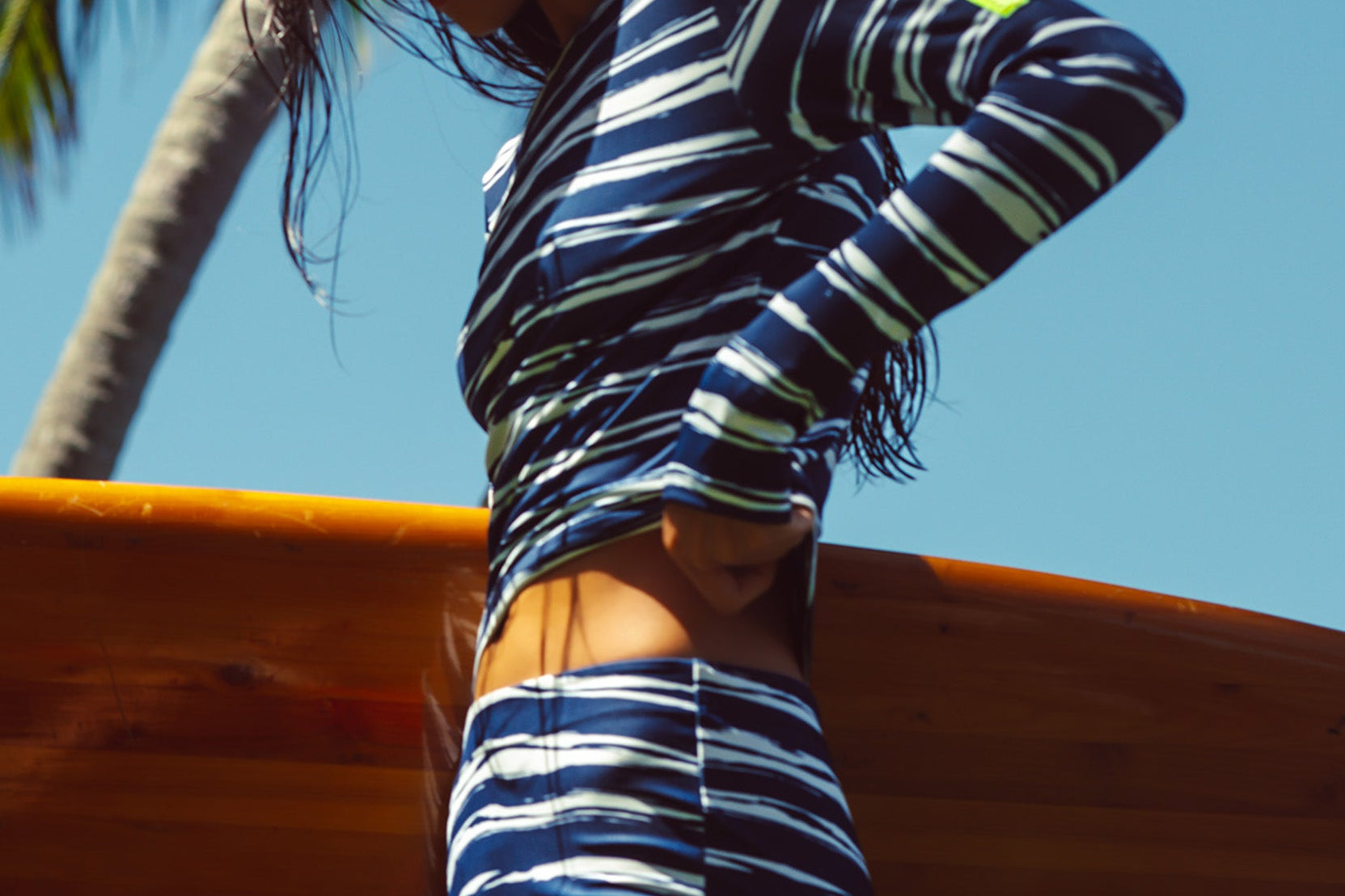 Woman wearing a wavy blue & white stripe tee and boy short holding a surf board in a tropical setting