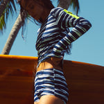 Woman wearing a wavy blue & white stripe tee and boy short holding a surf board in a tropical setting