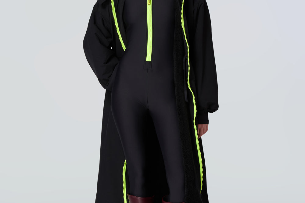 Front view of a woman wearing a black hooded long raincoat with yellow zipper and black catsuit with yellow zipper.
