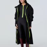 Front view of a woman wearing a black hooded long raincoat with yellow zipper and black catsuit with yellow zipper.