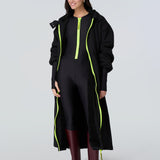 Front view of a woman wearing a black hooded long raincoat with yellow zipper and black catsuit with yellow zipper.
