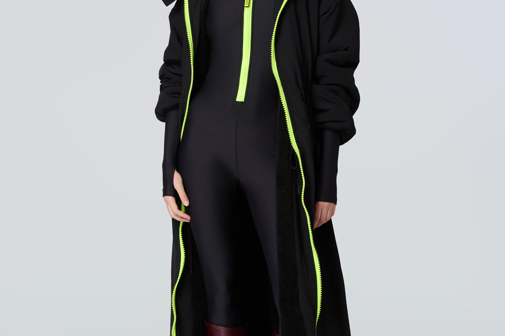 Front view of a woman wearing a black hooded long raincoat with yellow zipper and black catsuit with yellow zipper.