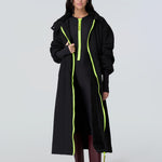 Front view of a woman wearing a black hooded long raincoat with yellow zipper and black catsuit with yellow zipper.