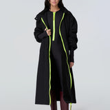 Front view of a woman wearing a black hooded long raincoat with yellow zipper and black catsuit with yellow zipper.