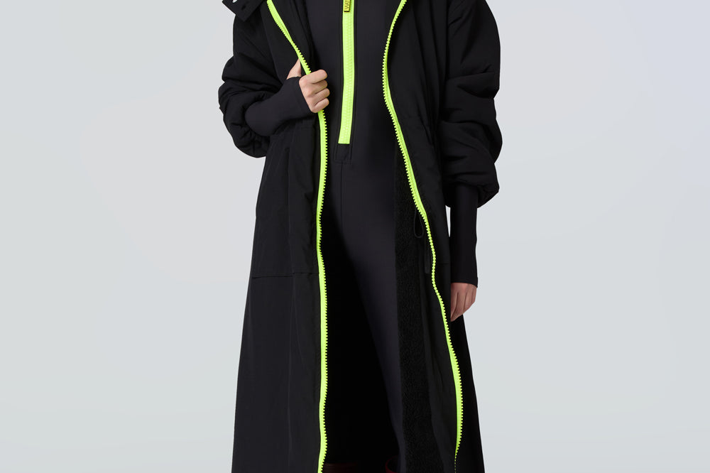 Front view of a woman wearing a black hooded long raincoat with yellow zipper and black catsuit with yellow zipper.
