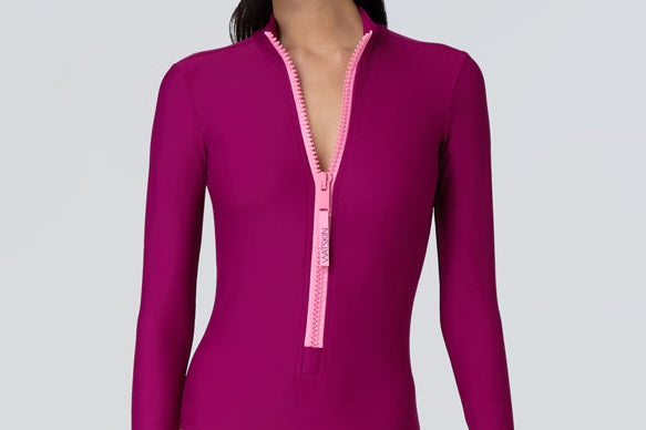 Front view of a woman wearing a long sleeve bodysuit in dark berry with a pink front zipper.