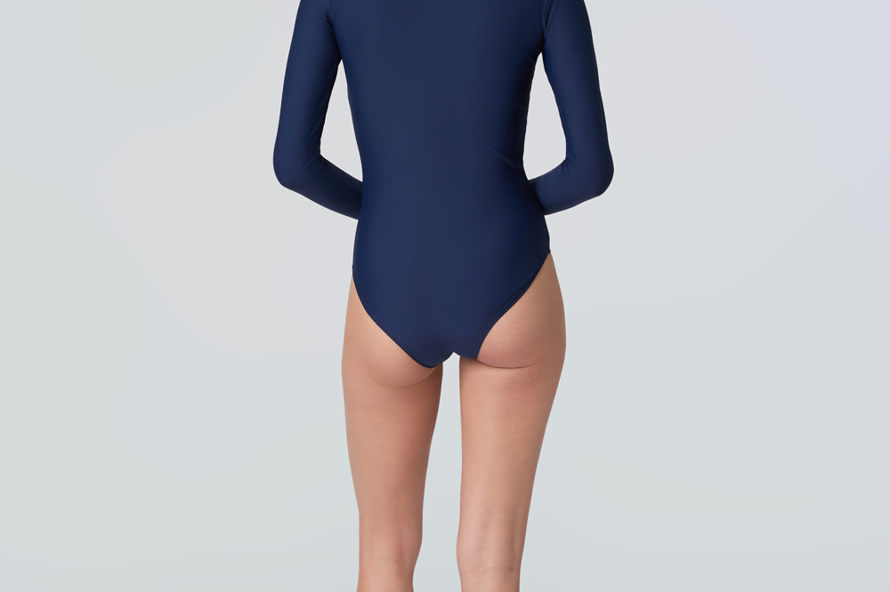 Back view of a woman wearing a long-sleeve navy blue one-piece swimsuit with a light blue front zipper.