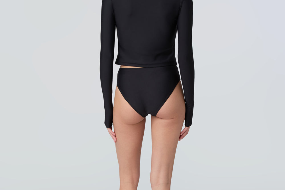 Back view of a woman wearing long sleeve mock neck zip front black rashguard with a yellow zipper and black bikini bottom.